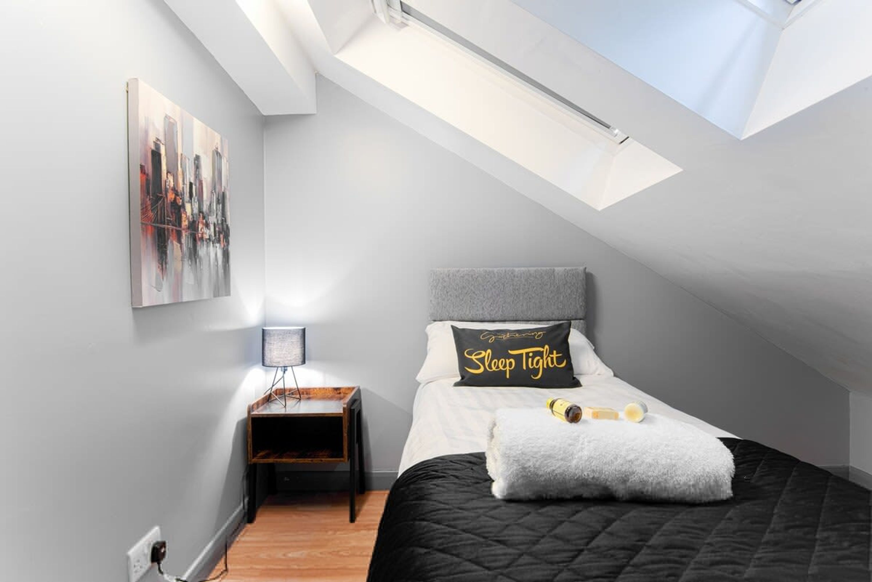 Un dormitorio con tragaluces, una cama con almohada, una mesita de noche y un cuadro colgado en la pared.