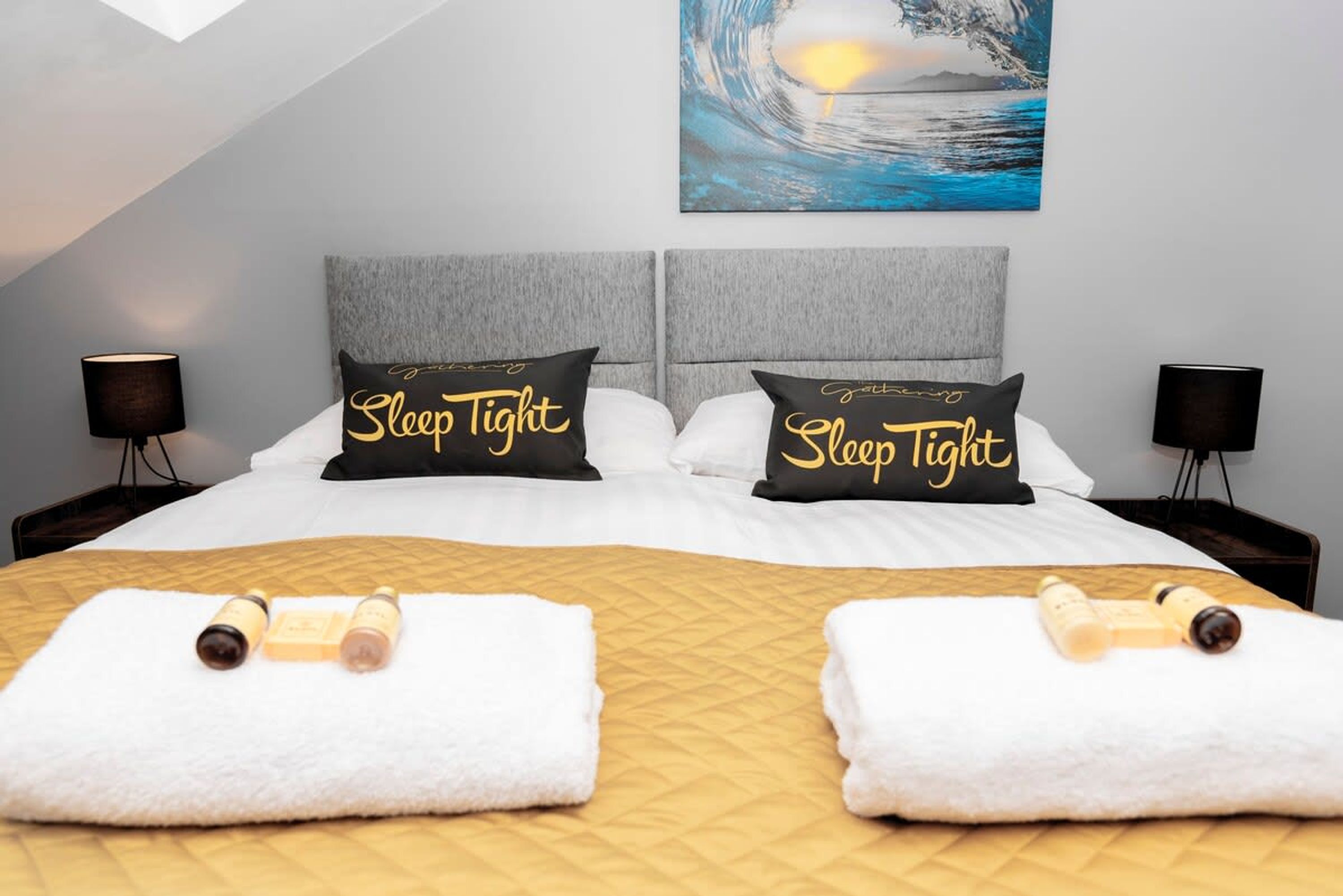 Una habitación de hotel con una cama, dos almohadas con el texto 'Sleep Tight', dos mesitas de noche, una lámpara, un cuadro de una ola y toallas con botellas.