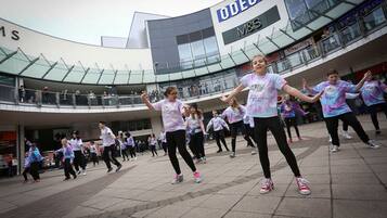 A group of people performing a dance routine in a public square with a modern building in the background.