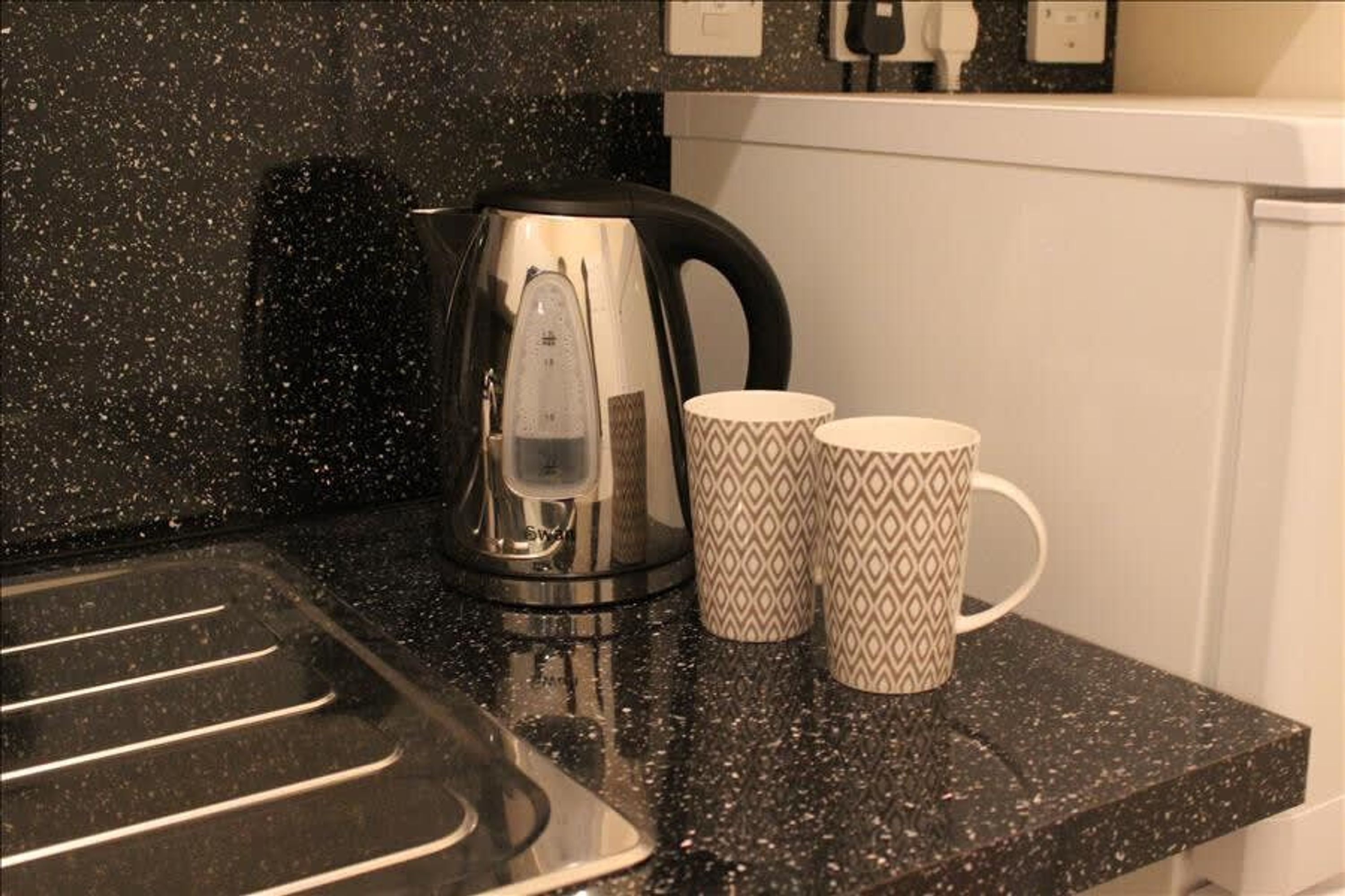 Una encimera de cocina con un hervidor de acero inoxidable, dos tazas estampadas y un salpicadero negro con salpicaduras.