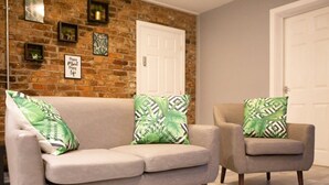 A living room with a brick wall, a white door, and a grey sofa and armchair with green patterned cushions. - The Mersey Apartment Liver House (Birkenhead)