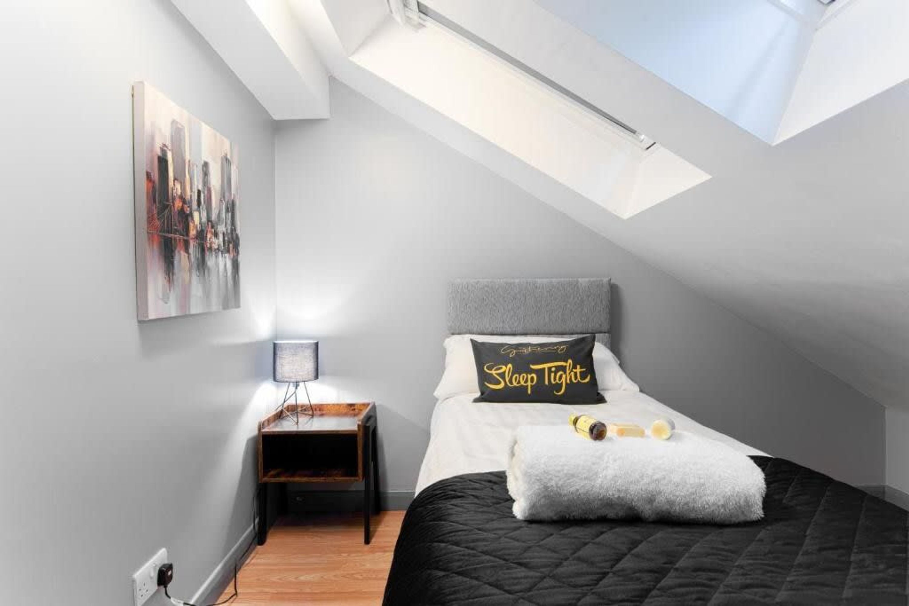 Una camera da letto con un lucernario, un letto con un cuscino con la scritta 'Sleep Tight', un comodino con una lampada e un quadro raffigurante un panorama urbano.