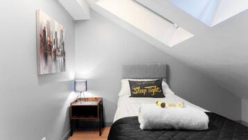 A bedroom with a skylight, a bed with a 'Sleep Tight' pillow, a bedside table with a lamp, and a cityscape painting on the wall.
