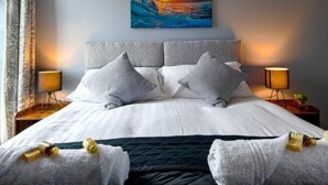 A modern hotel room with a large bed, grey bedding, and a painting of a sunset over the ocean on the wall.