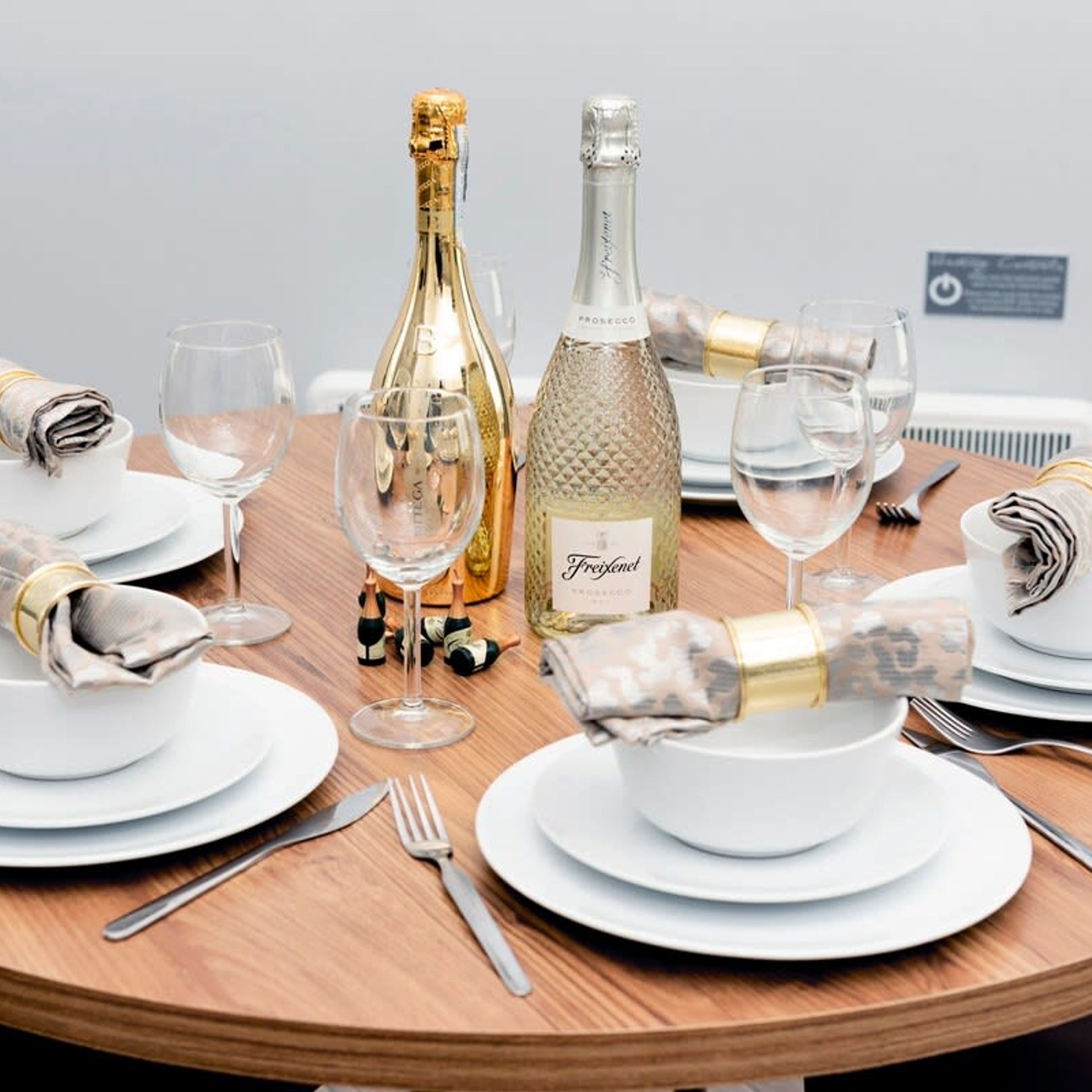 A round dining table set with white plates, glasses, and cutlery, with two bottles of champagne and a folded napkin.