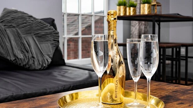 A room with a large window, a black sofa, a wooden table with a gold tray holding a champagne bottle and two glasses. - The Maelor - Berwyn House Central Wrexham (Wrexham)