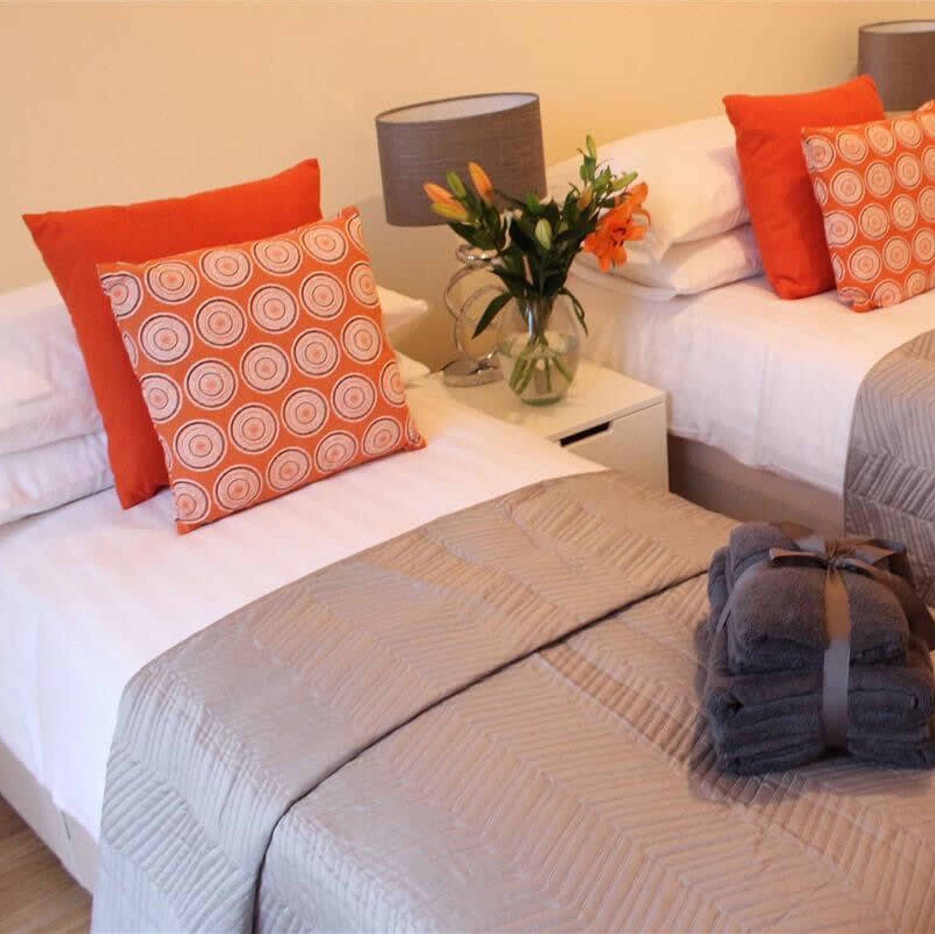 A hotel room with two beds, each with white bedding and orange patterned pillows. There is a vase with flowers on the nightstand between the beds.