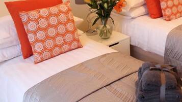 A hotel room with two beds, each with white bedding and orange patterned pillows. There is a vase with flowers on the nightstand between the beds.