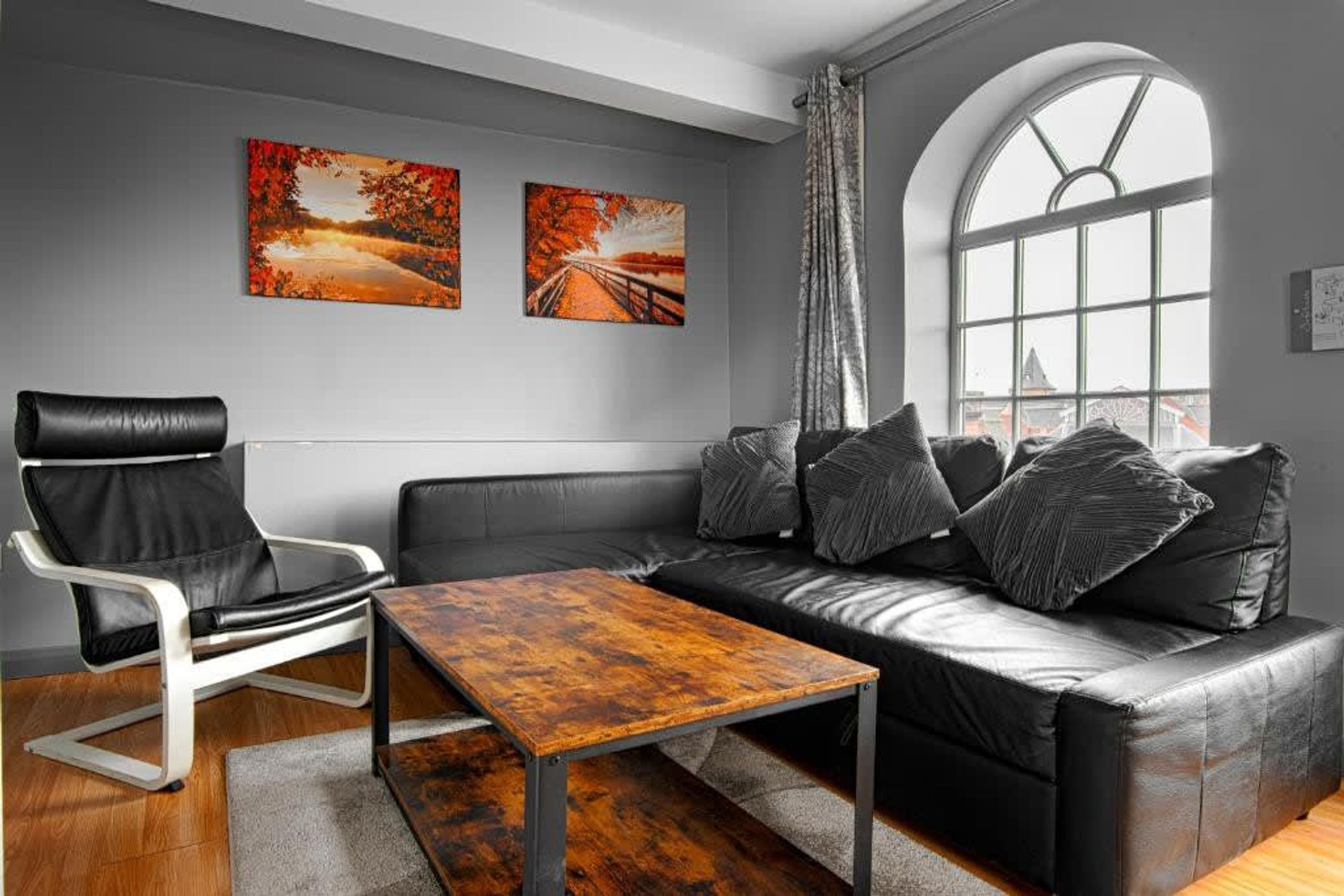 Una sala de estar moderna con un sofá de cuero negro, una silla blanca y una mesa de centro de madera. Hay dos cuadros en la pared, una ventana con vistas y una cortina.