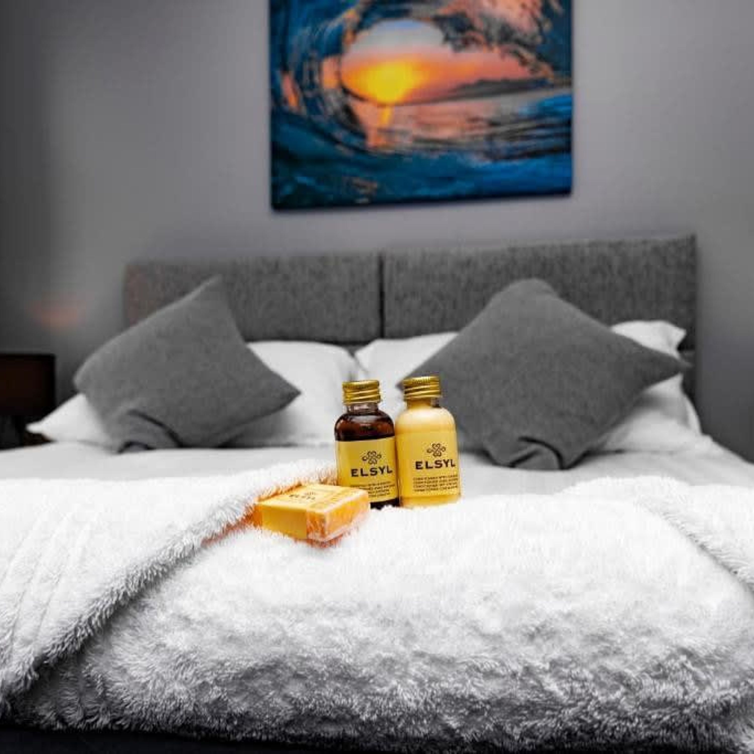 Uma cama bem arrumada com um cobertor branco e fofo, duas embalagens de produtos de banho ELSYL e uma barra de sabão sobre um travesseiro cinzento.
