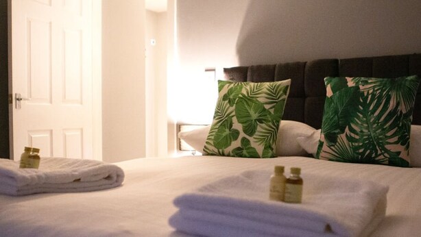 Una habitación de hotel con una cama bien hecha, dos almohadas con estampado de hojas tropicales y una puerta visible.