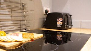Una cocina con una placa de inducción negra, una tabla de madera con pan y queso rebanados, y un tostador negro sobre una encimera de madera.