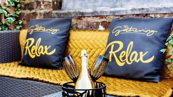 Eine entspannte Sitzgelegenheit im Freien mit einer Champagnerflasche und zwei Gläsern, ergänzt durch Kissen mit dem Aufdruck 'The Gathering Relax'.