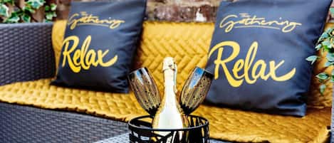 A relaxed outdoor seating area with a bottle of champagne and two glasses, accompanied by pillows with the text "The Gathering Relax".