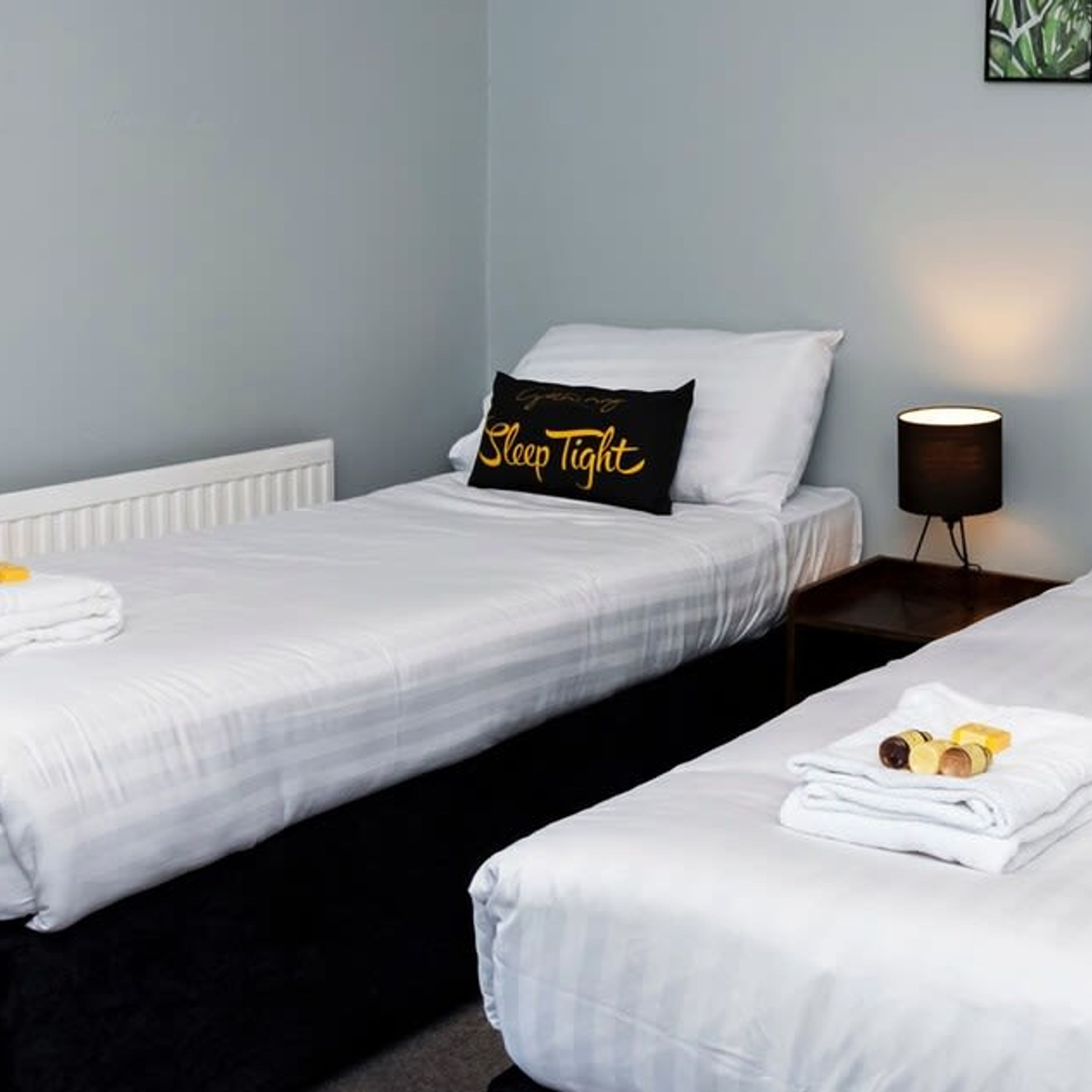 A hotel room with two single beds, a bedside table with a lamp, and a pillow with the text "Sleep Tight".