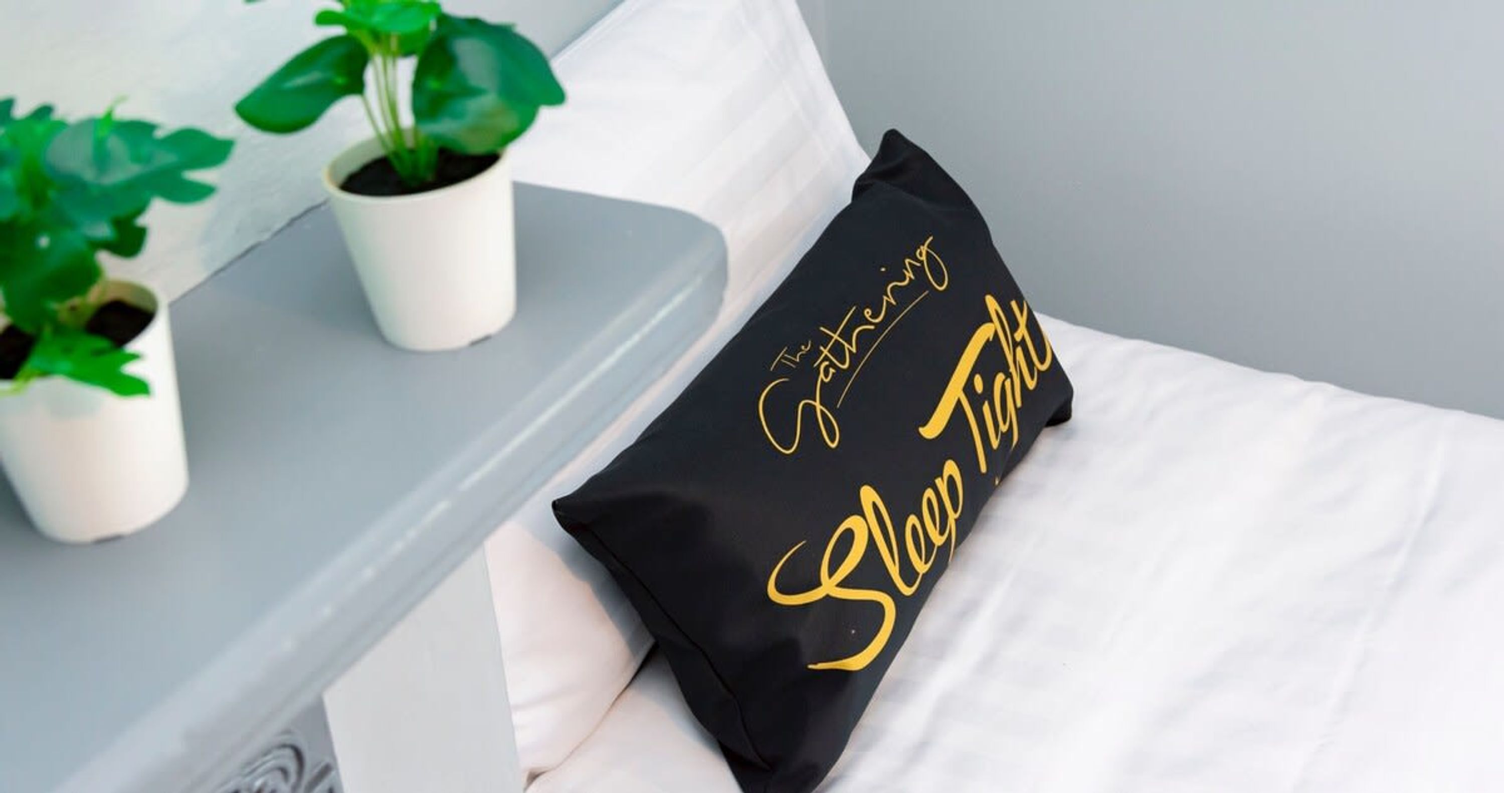 Una cama con ropa blanca y un almohadón negro con letras doradas. A la izquierda, dos macetas sobre una superficie gris.
