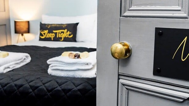 Una habitación de hotel con una cama, un almohadón con el texto 'Sleep Tight', un letrero en la pared con la letra 'N' y una puerta con picaporte de latón.