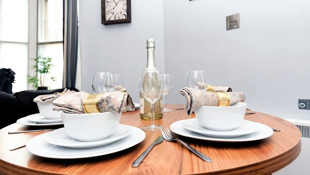 A dining table set for two with white plates, bowls, and cutlery, a bottle of champagne, and a clock on the wall. - The Lower Gathering - Wrexham Town Centre - Sleeps Up To 10 (Wrexham)