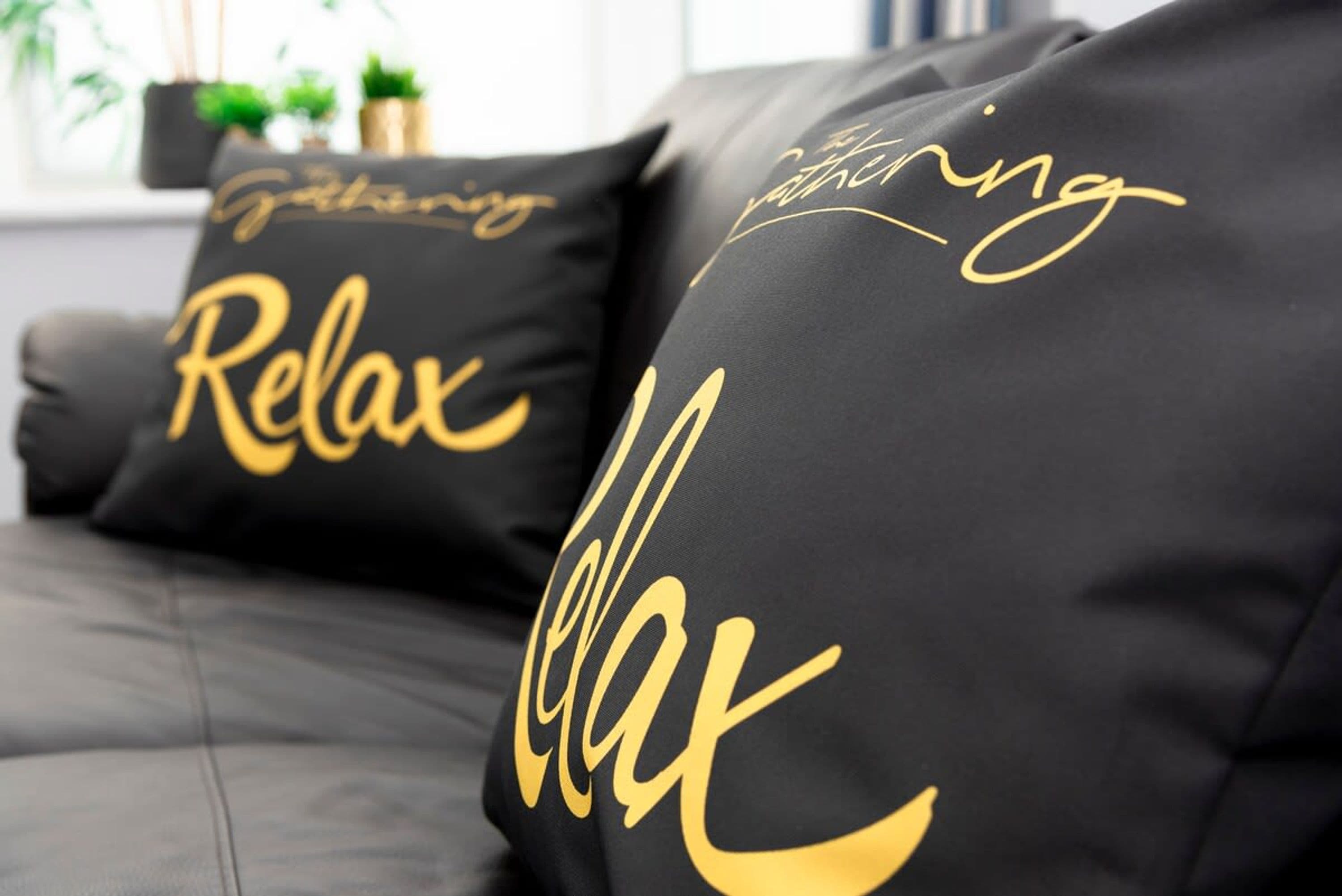 A black sofa with two cushions featuring the word "Relax" in gold lettering.