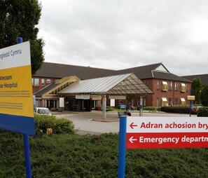 A hospital entrance with a covered waiting area and a sign indicating directions to the emergency department. - Wrexham Central Stay Apartment Seven (Wrexham)