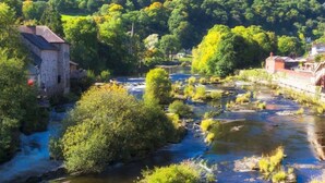 A riverside building with a stone facade, surrounded by lush greenery and a forested hill in the background. - Wrexham Central Stay Apartment Seven (Wrexham)