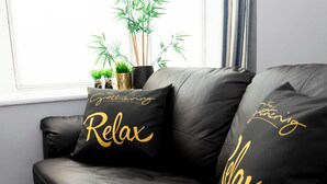 A black leather sofa with two cushions, one with "The Gathering" and "Relax" text, and another with "The Gathering" and "Calm" text. - Wrexham Central Stay Apartment Seven (Wrexham)