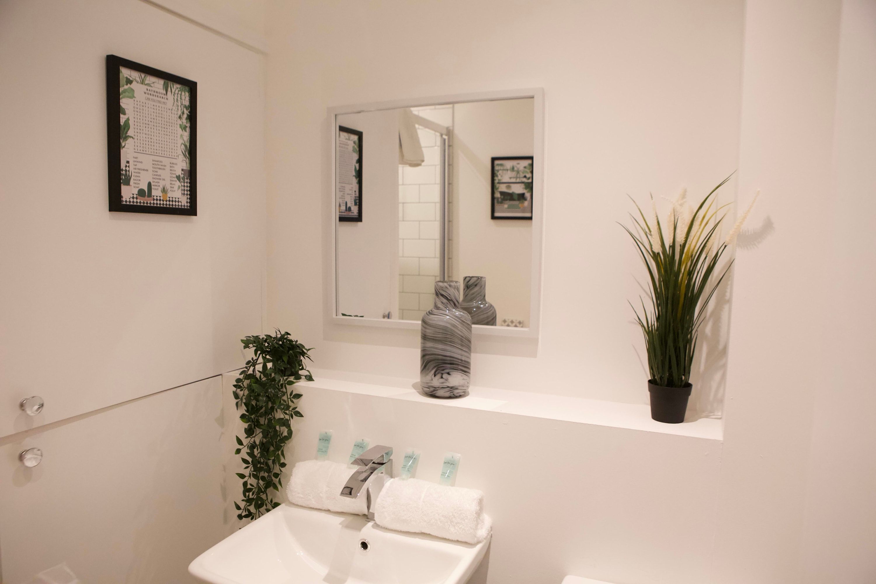 Een badkamer met een witte wastafel, een spiegel, een potplant en een lijst met een afbeelding aan de muur.