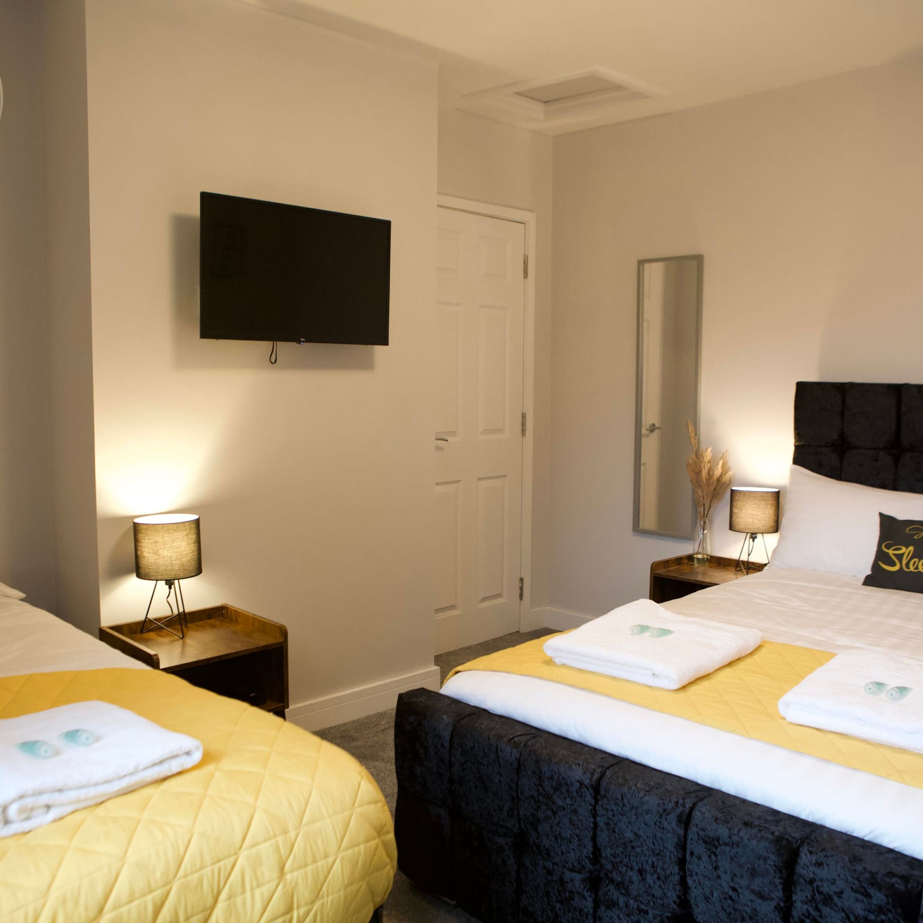 A hotel room with two beds, a flat-screen TV mounted on the wall, a mirror, and a bedside table with a lamp.