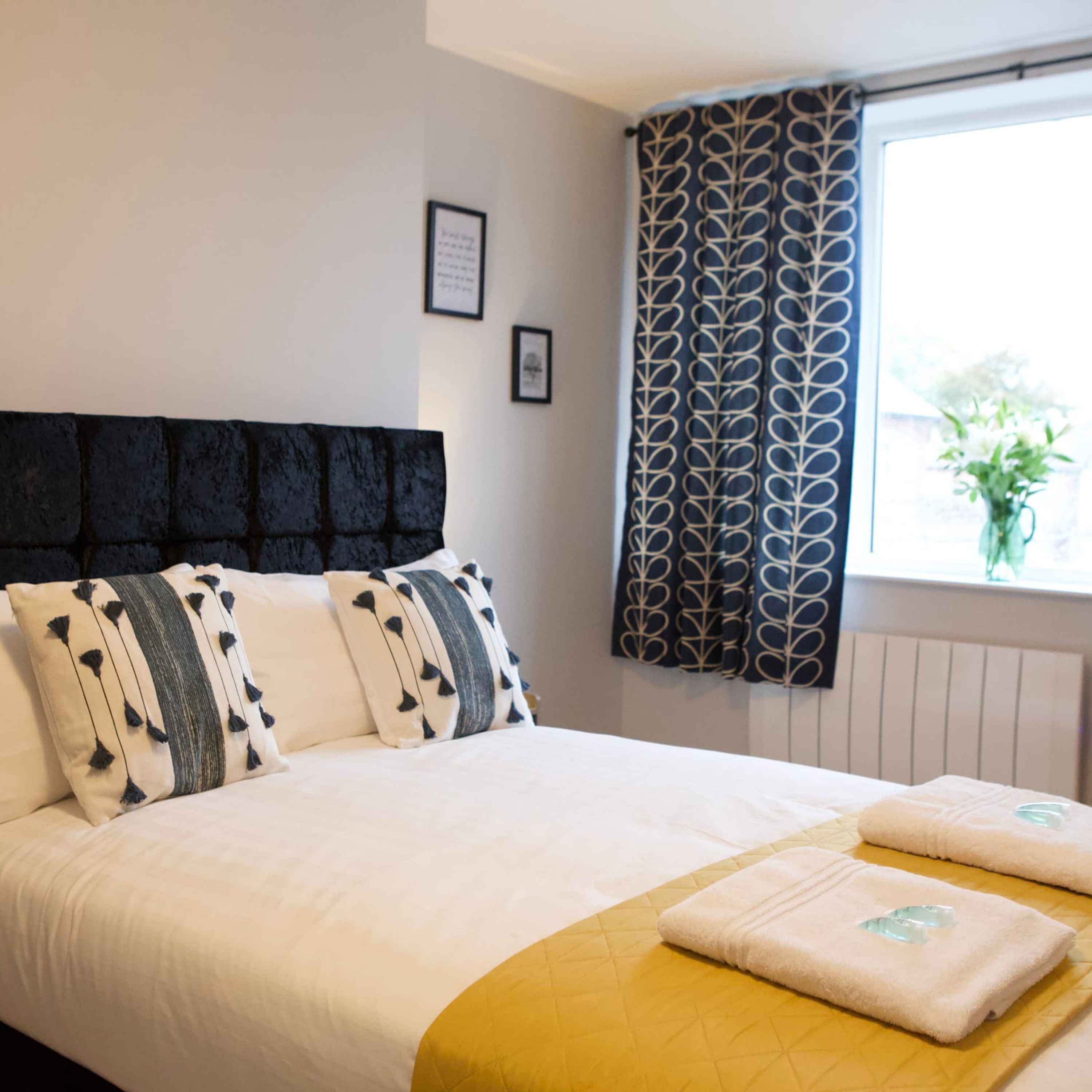 A neatly made bed with a headboard, a window with patterned curtains, and a bedside table with towels.