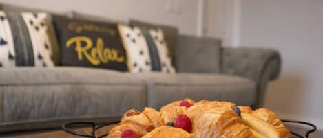A tray of croissants and berries on a brown table in a living room with a grey sofa and a 'Relax' pillow.