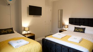A hotel room with two beds, each with a 'Sleep Tight' pillow, a flat-screen TV mounted on the wall, and a standing fan.
