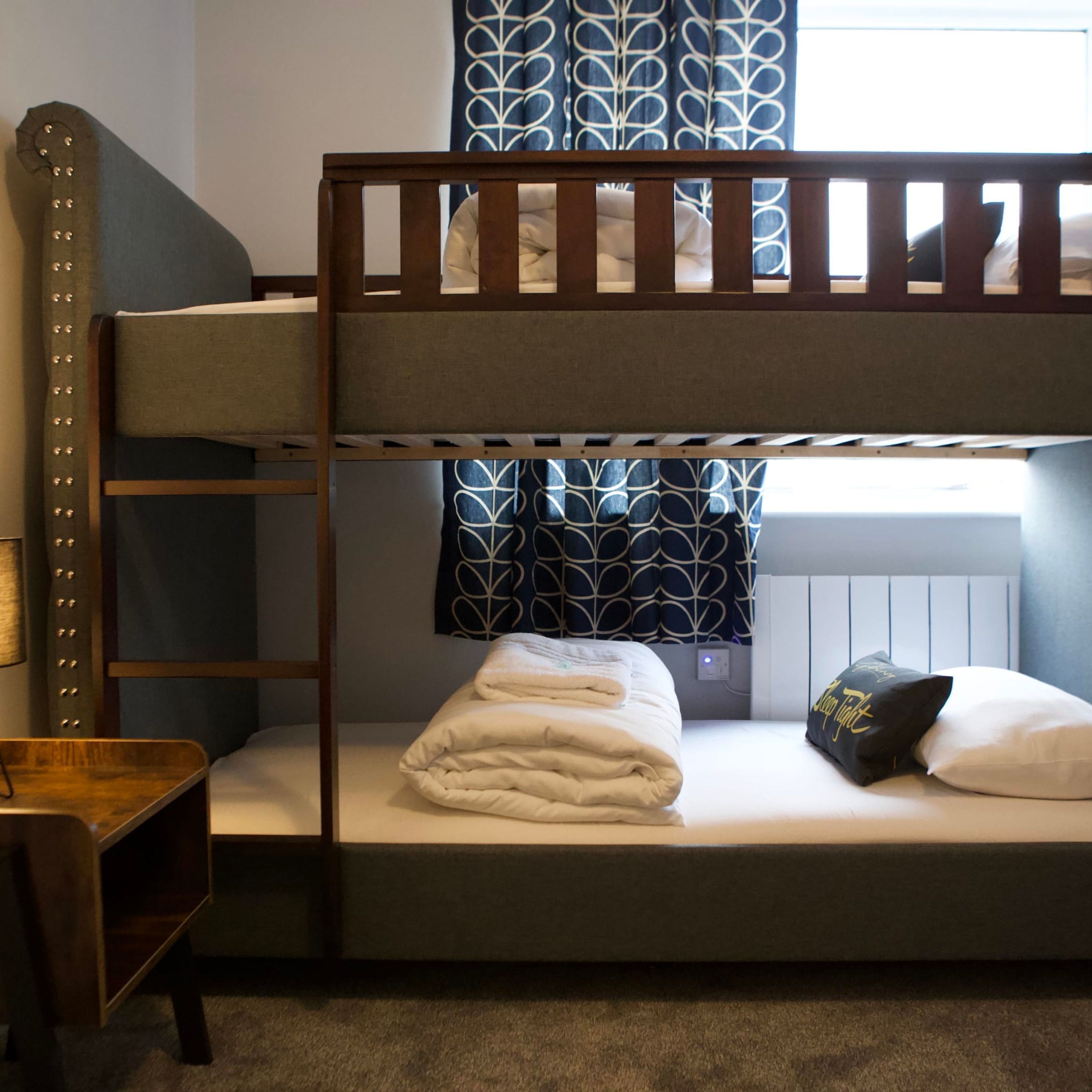 A bunk bed with a wooden frame, a bedside table, a lamp, and a window with patterned curtains.