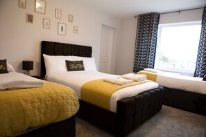 Two single beds with dark headboards and white bedding, a large window with an outside view, and framed artwork adorn the walls.