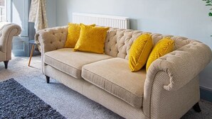 Ein beiges Sofa mit gelben Kissen, ein weißer Heizkörper und gerahmte Pflanzenbilder an der Wand.