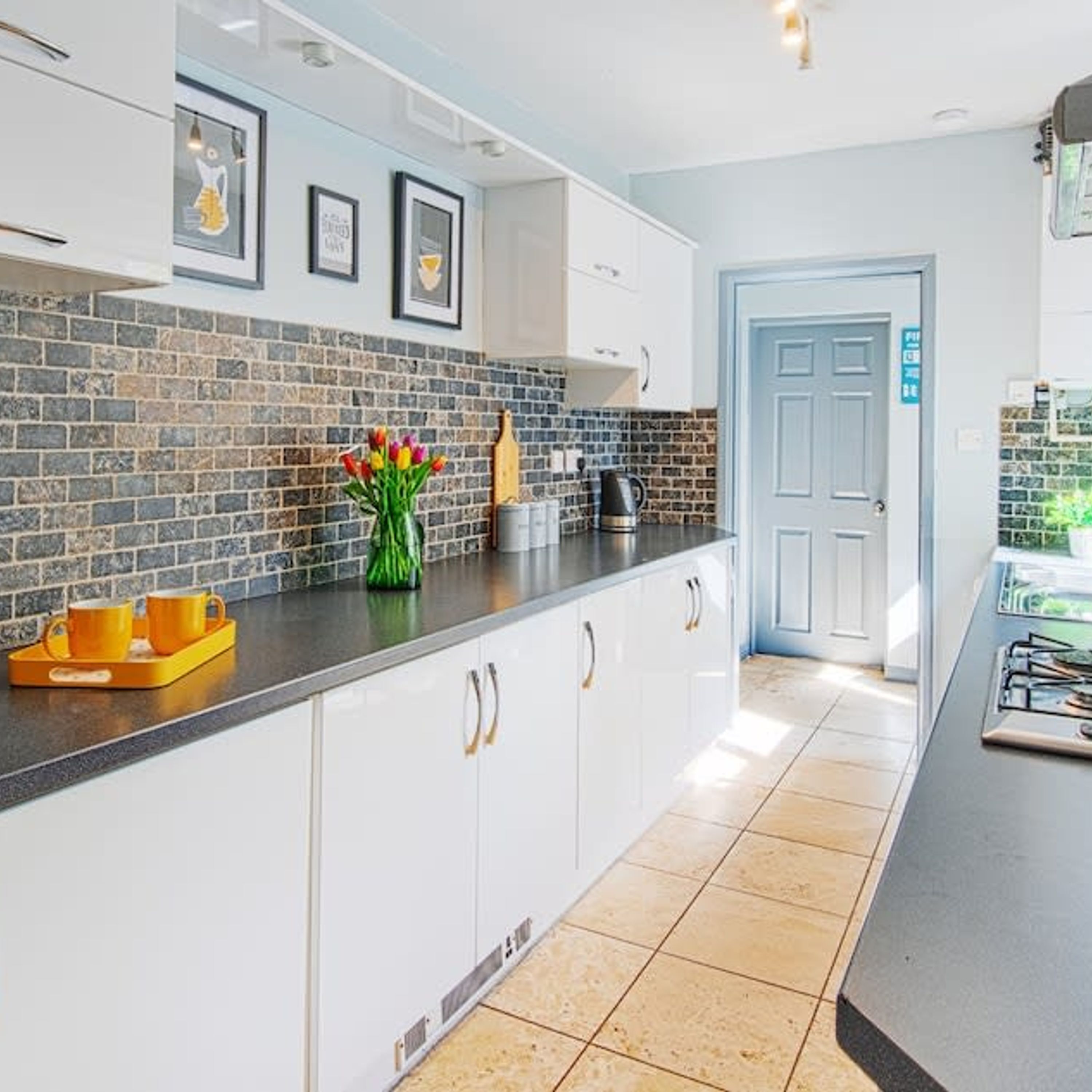 A modern kitchen with white cabinets, a black countertop, and a tiled backsplash. There is a coffee maker, a kettle, and a tray with orange cups.