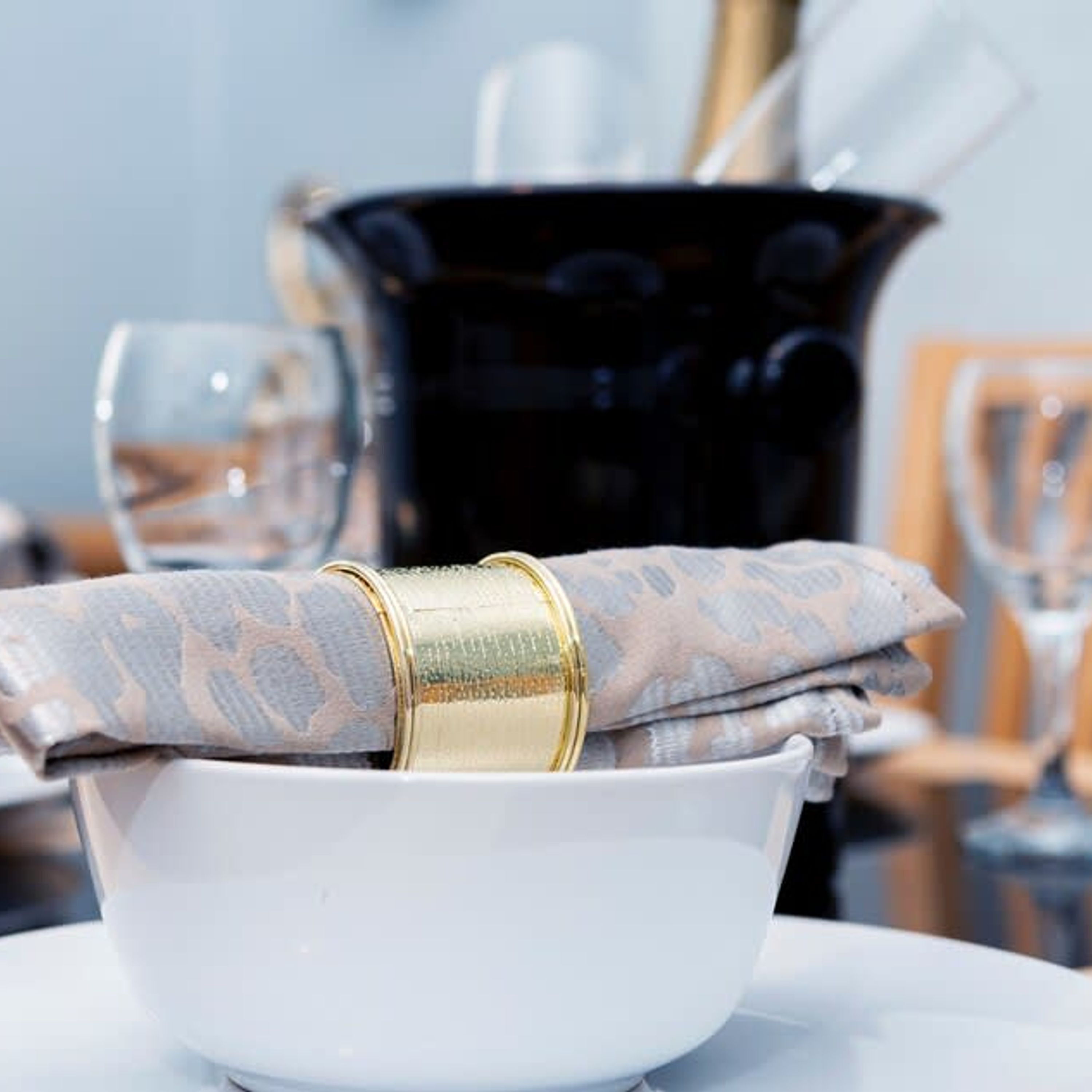 A white bowl with a patterned napkin and a gold napkin ring on a table.