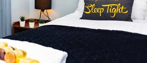 Una cama bien hecha con edredón blanco y negro, una almohada con el texto "Sleep Tight" y una mesita de noche con una lámpara y pequeñas plantas.