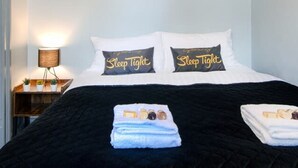 Ein ordentlich bezogenes Bett mit einer schwarzen Tagesdecke, zwei Kissen mit dem Aufdruck 'Sleep Tight' und ein Nachttisch mit Lampe und kleiner Pflanze.