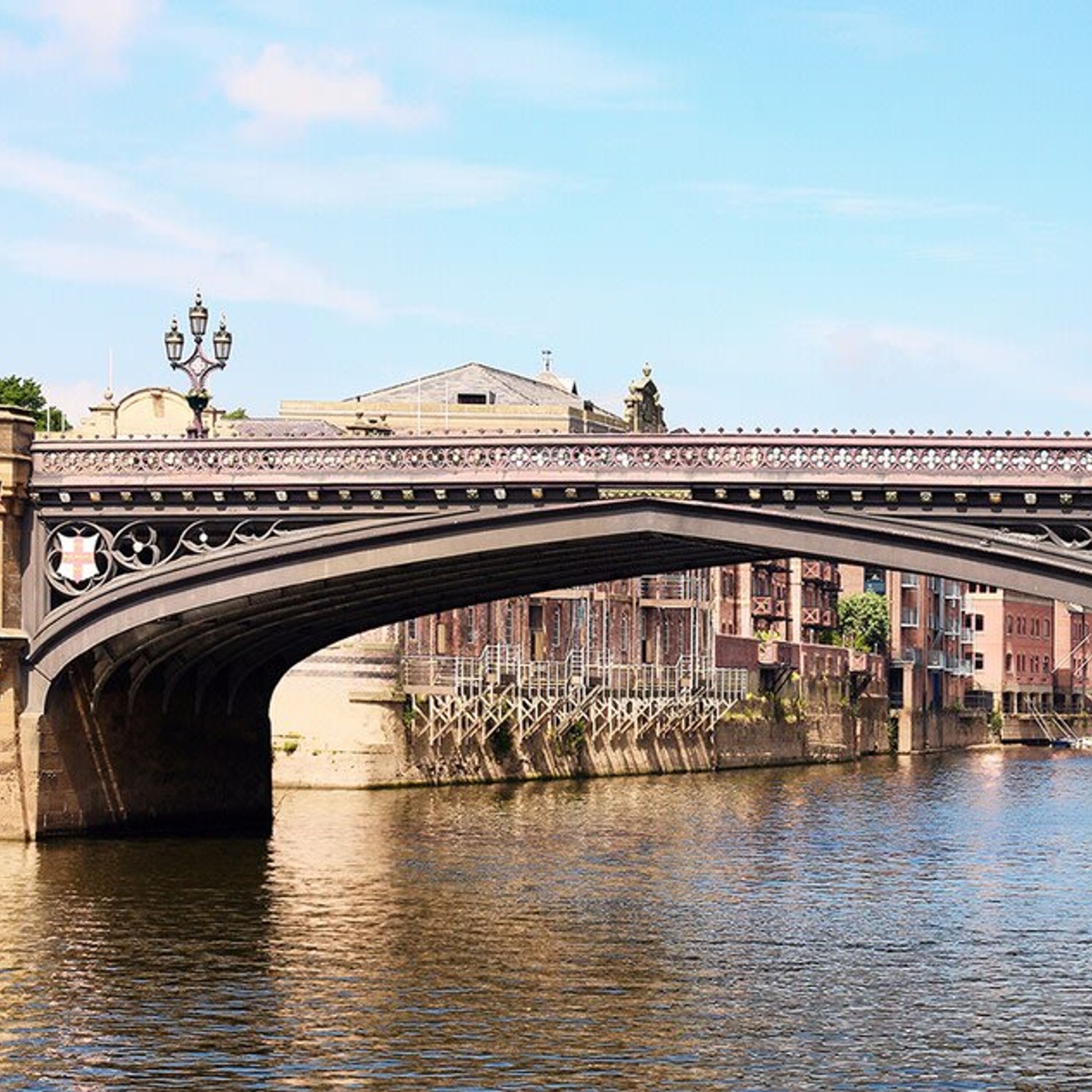 A stone bridge with ornate railings spanning over a river, with buildings and a lamp post in the background.