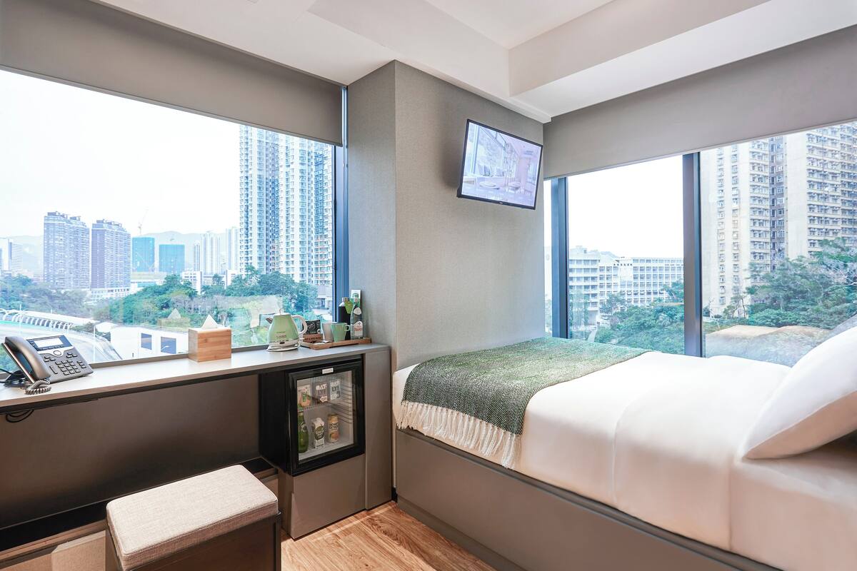 Une chambre d’hôtel compacte comprenant un lit, un bureau avec un ordinateur, un mini-réfrigérateur et une fenêtre donnant sur la ville.