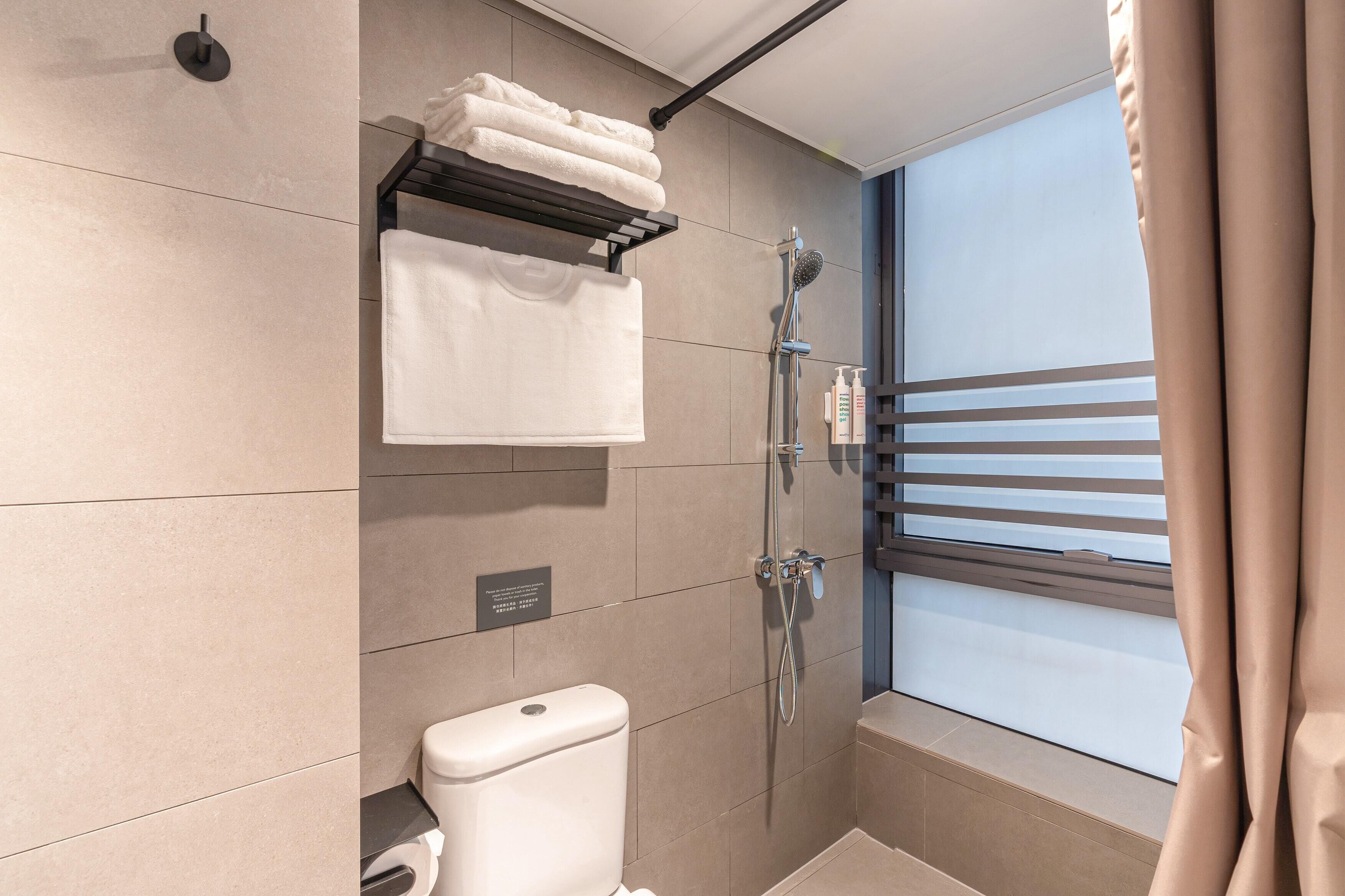 A modern bathroom with a toilet, a towel rack with folded towels, a shower with a handheld showerhead, and a window with blinds.