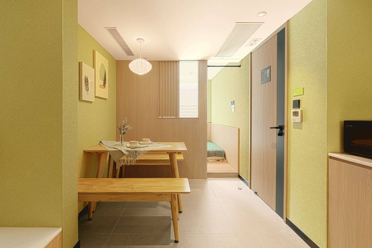 Un couloir avec un banc en bois, une table et un lit. Des tableaux encadrés ornent le mur, il y a une fenêtre avec des stores et une porte sur laquelle est apposée une pancarte.