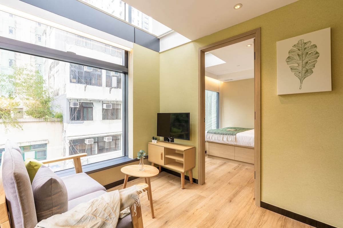Une chambre d’hôtel moderne dotée d’une grande fenêtre, d’un téléviseur à écran plat, d’une armoire en bois, d’une petite table ronde et d’un lit recouvert d’une couverture verte.