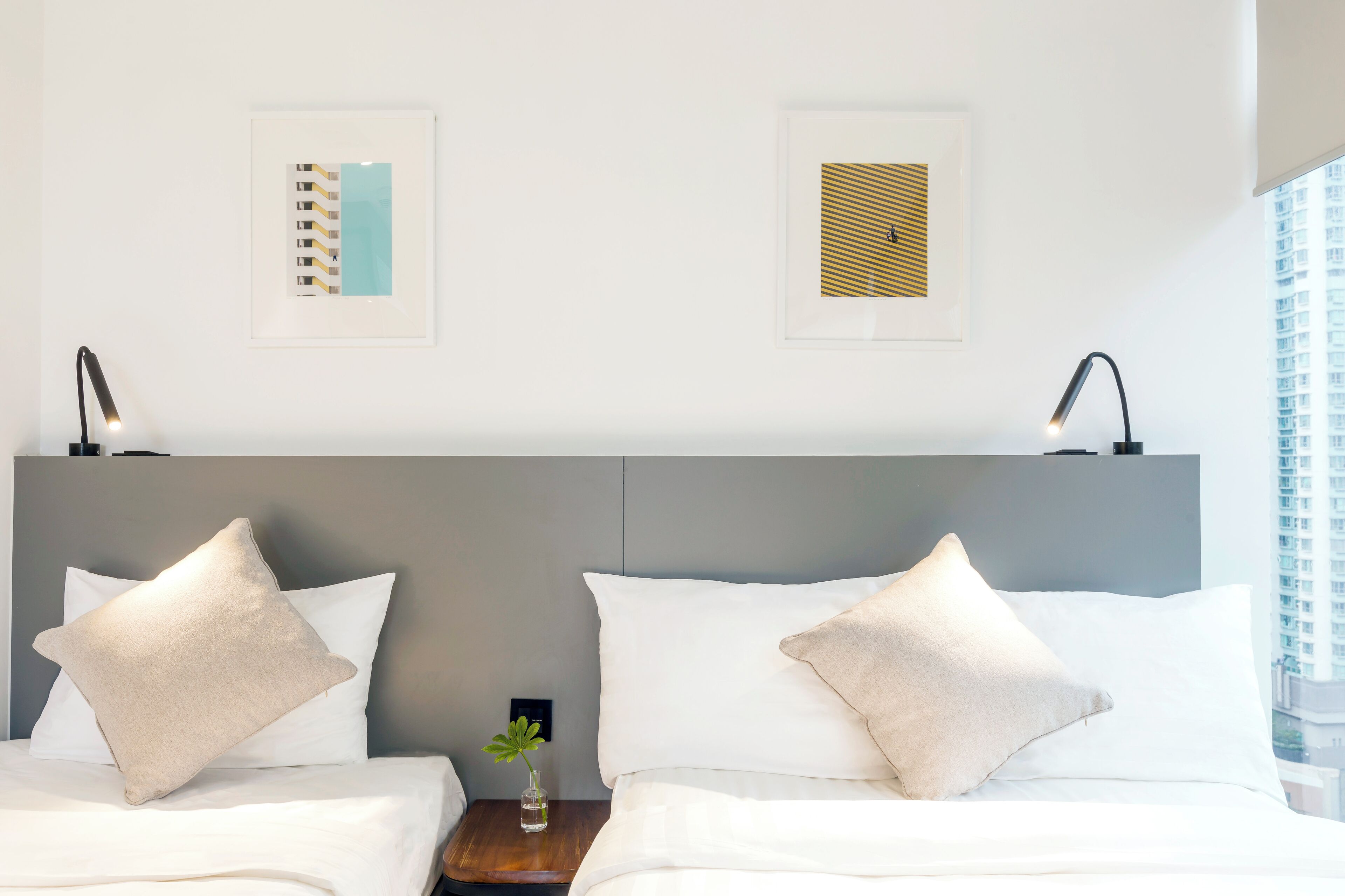 A hotel room with two beds, a grey headboard, framed artwork on the wall, and a small bedside table with a lamp.