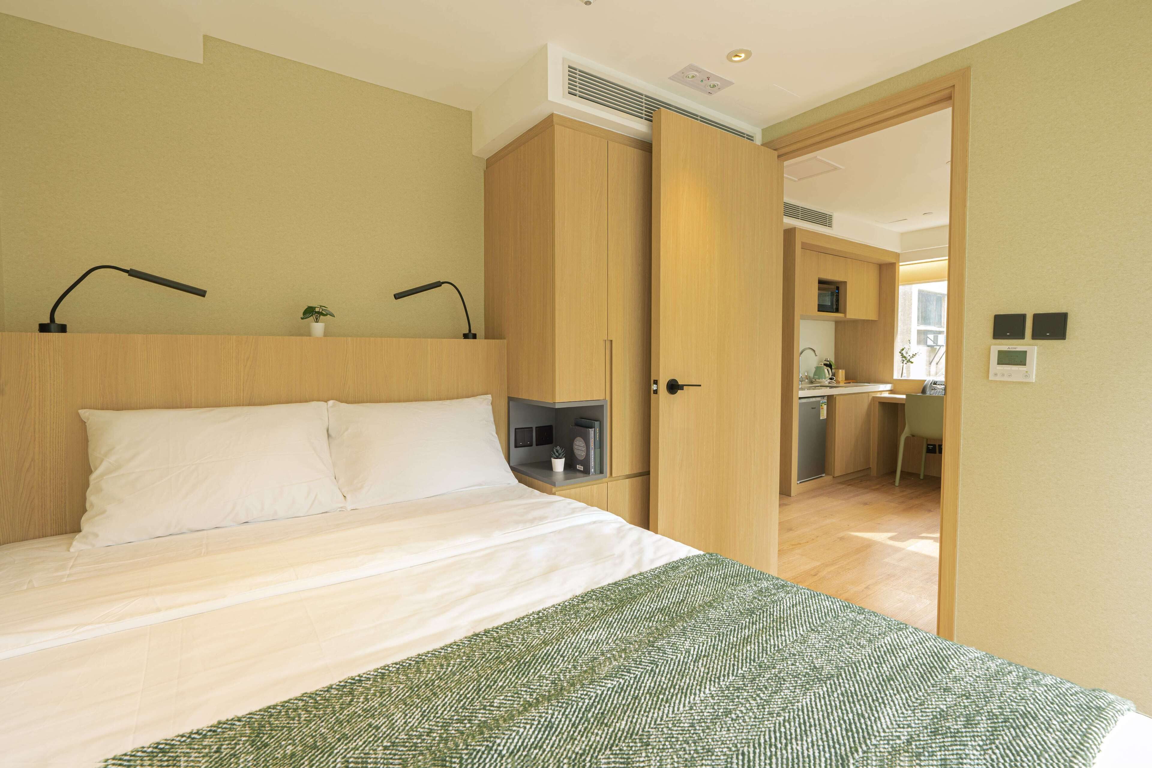 A bedroom with a bed, bedside lamps, a green patterned throw, wooden wardrobe, and an open door leading to a kitchen area.