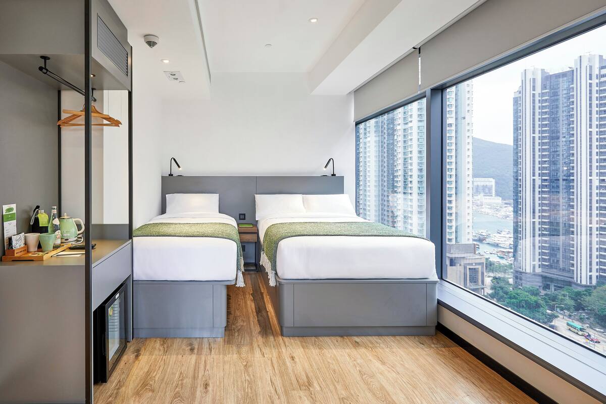Une chambre d’hôtel moderne avec deux lits, une grande fenêtre donnant sur la ville et un petit bureau équipé d’un miroir et de produits de toilette.