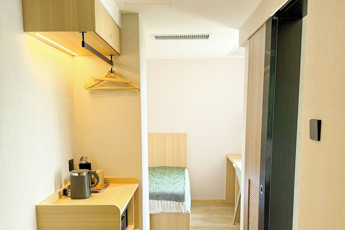 A narrow hallway with a kitchenette on the left, a bed with a green blanket in the center, and a black door on the right.