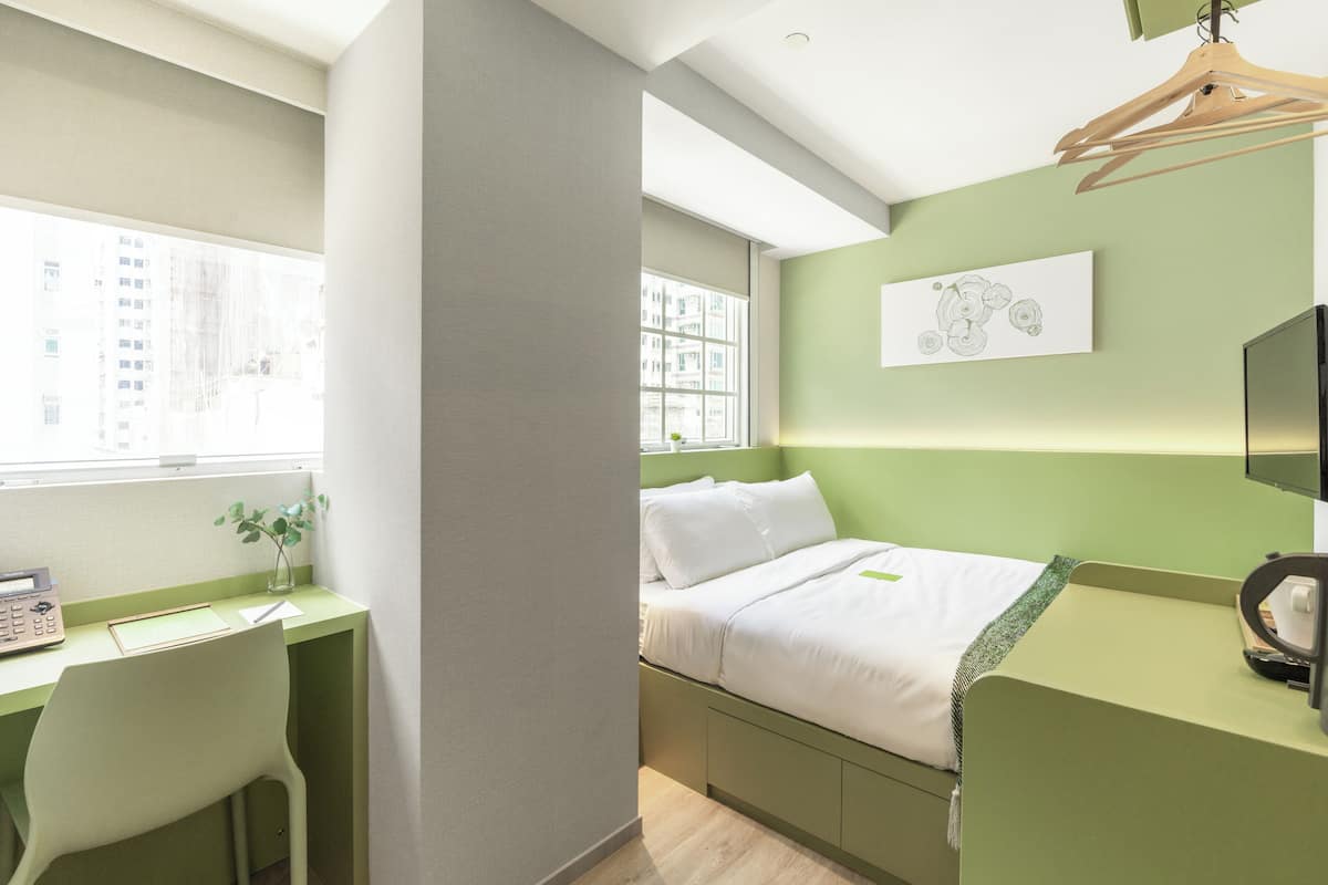A modern bedroom with a green accent wall, a bed with white bedding, a desk with a chair, and a window with a view of buildings.