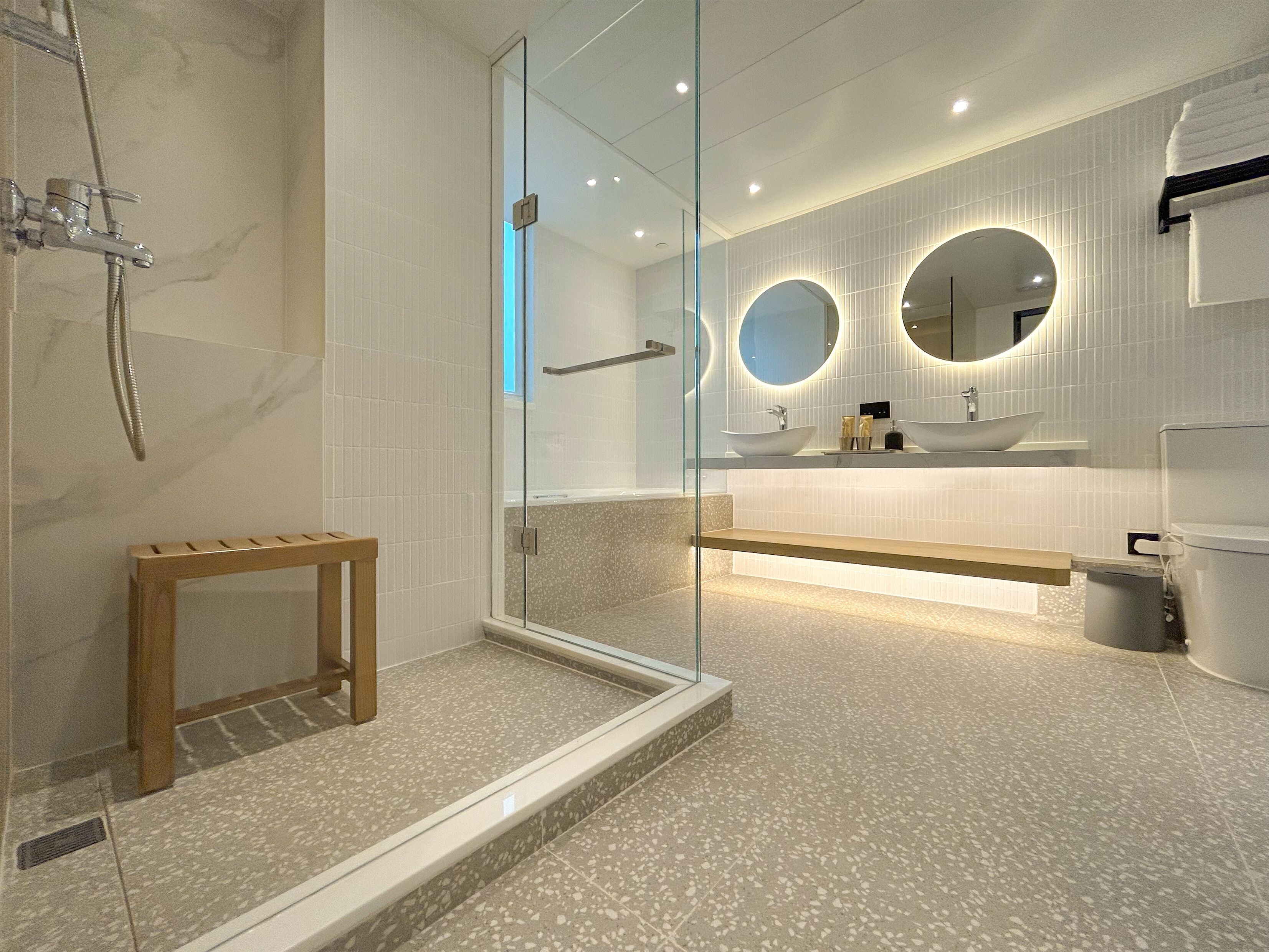 A modern bathroom with a glass-enclosed shower, a wooden bench, and a vanity with two round mirrors.