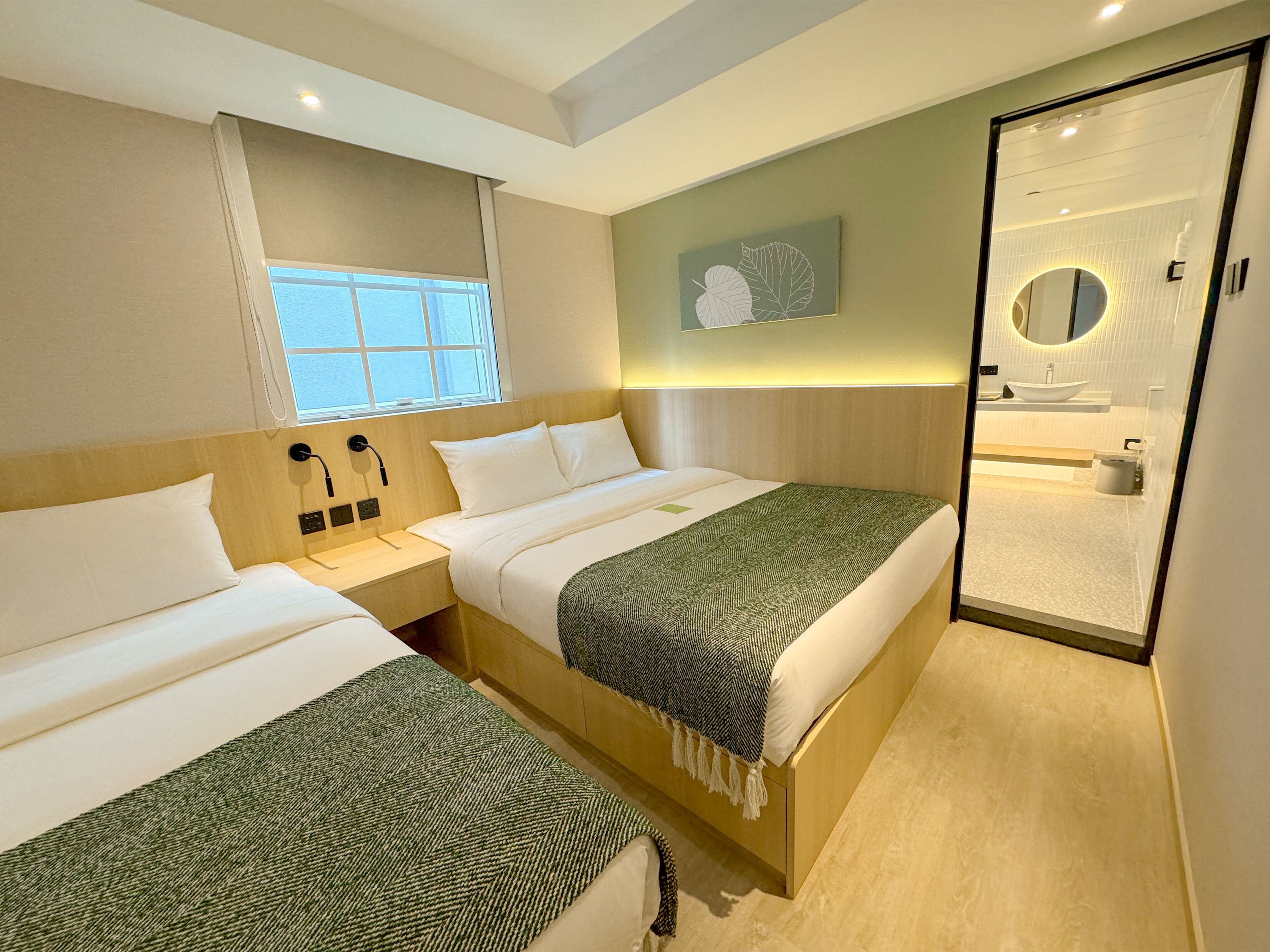 Une chambre d’hôtel avec deux lits, une grande fenêtre, une œuvre d’art accrochée au mur et une salle de bain visible à travers une porte en verre.